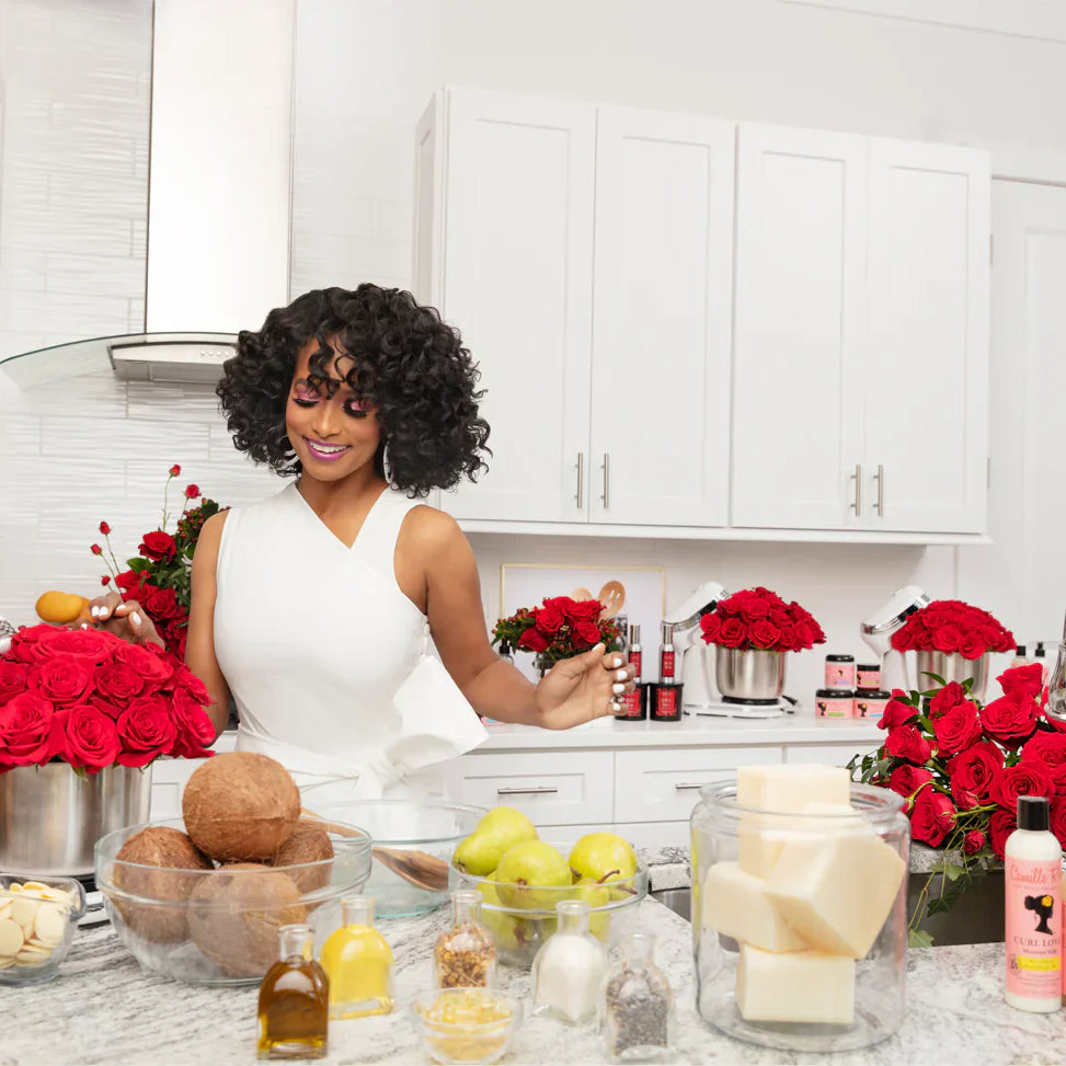 Vrouw met krullend haar in een keuken omringd door natuurlijke ingrediënten zoals kokos, oliën en verse bloemen, terwijl Camille Rose haarproducten op het aanrecht staan.