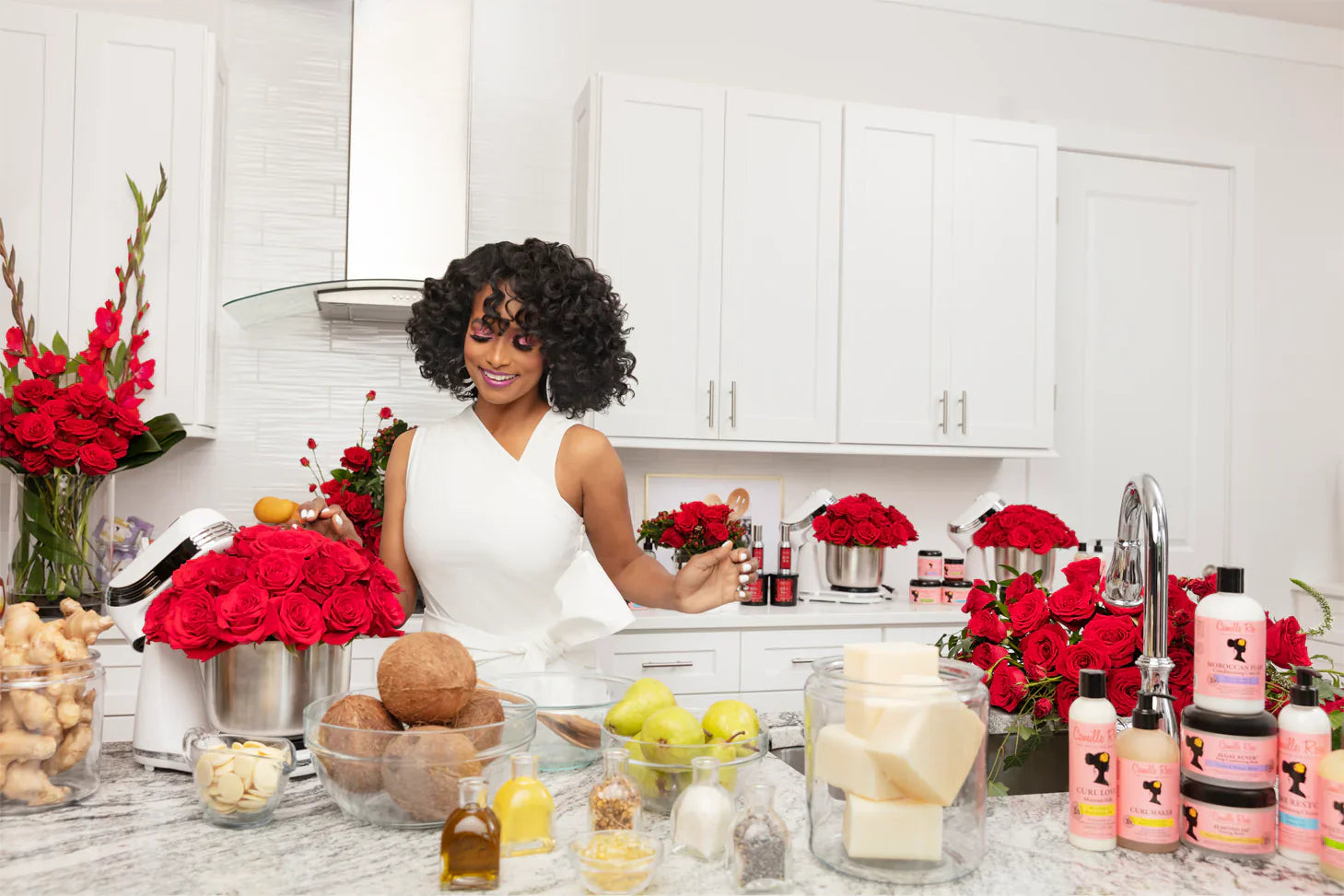 Vrouw met krullend haar in een keuken omringd door natuurlijke ingrediënten zoals kokos, oliën en verse bloemen, terwijl Camille Rose haarproducten op het aanrecht staan.