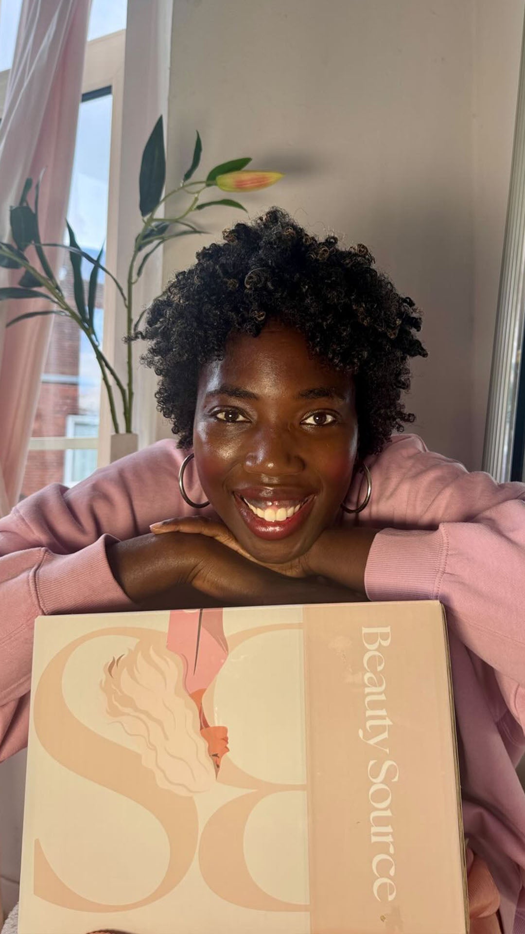 Michelle Amo met natuurlijk krullend haar glimlacht en houdt een Beauty Source box vast – ontdek verzorgingsproducten en haarverzorging online kopen bij Beauty Source.