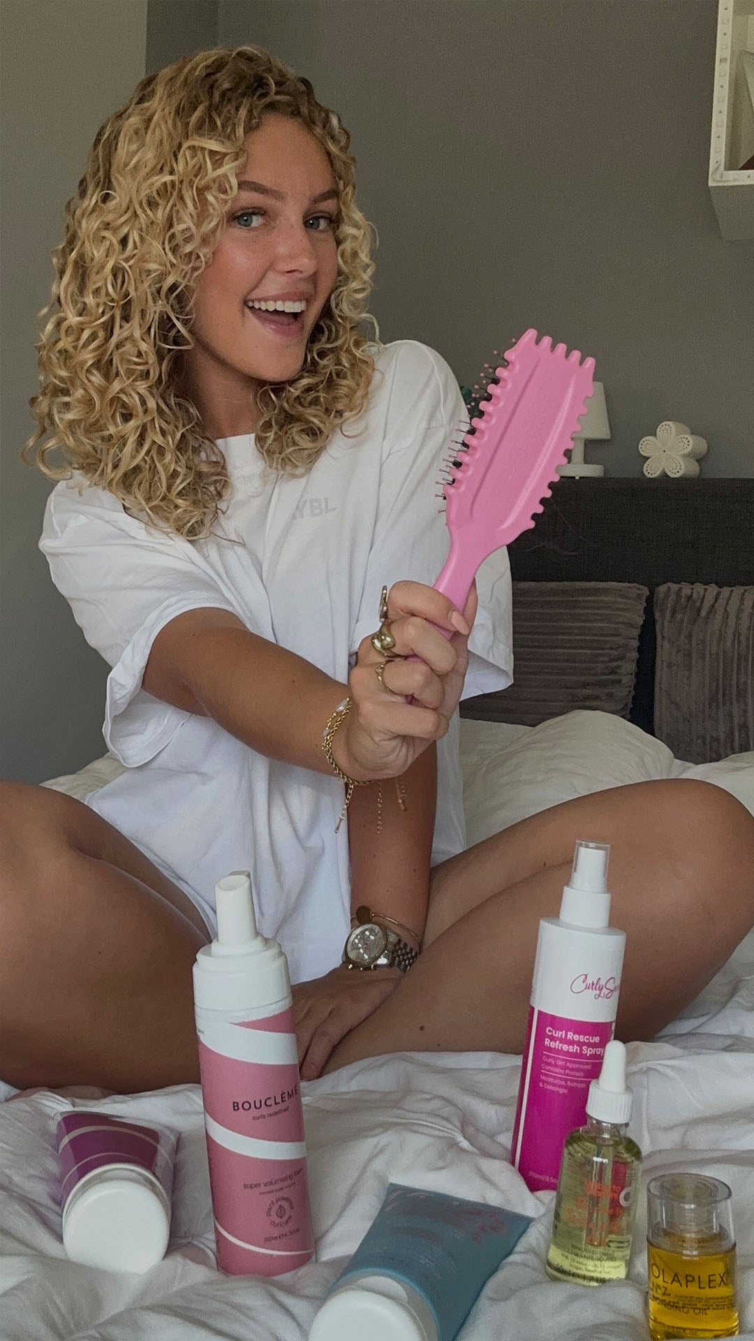 Kelly.vdp met blonde krullen zit op bed met haarborstel en haarverzorgingsproducten van Bouclème, Olaplex en Curly Secret – producten voor krullend haar online kopen bij Beauty Source.